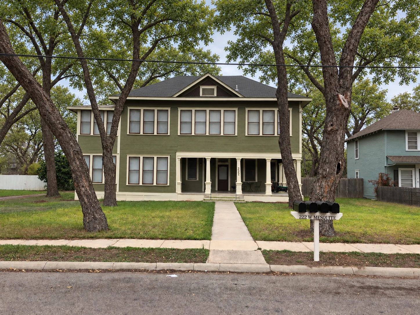 Front elevation of Mesquite House showing its olive-green exterior, cream trim, front porch, and mature trees